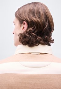 Άτομο με μεσαίου μήκους κυματιστά καστανά μαλλιά, ορατό από πίσω, φοράει πουκάμισο με γιακά σε μπεζ και λευκό χρώμα, με φόντο απλό και ανοιχτόχρωμο.