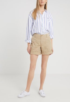 Kobieta ubrana w beżowe szorty z podwiniętymi nogawkami, białe trampki oraz białą koszulę z długim rękawem w niebieskie pionowe paski, stojąca na jednolitym białym tle.