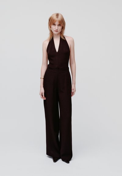Giovane donna con capelli biondo chiaro che indossa un jumpsuit senza maniche nero con collo all'americana e scarpe nere a punta, in piedi su uno sfondo bianco uniforme.