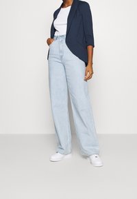 Marinblazer med en flödande design, i kombination med högmidjade, ljusblåvida byxor och vita sneakers. Enkel, modern klädsel.