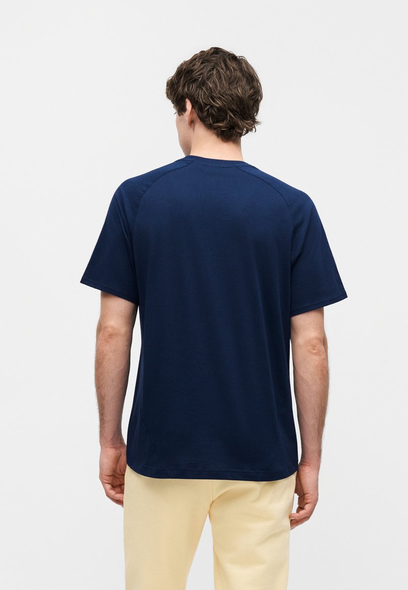 Man with curly brown hair wearing a plain navy blue short-sleeve shirt and light beige pants, standing facing away on a white background.