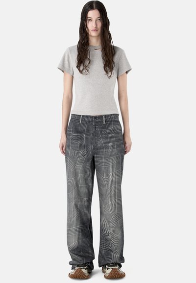 Mujer con el pelo largo y mojado lleva una camiseta gris de manga corta, pantalones sueltos a cuadros blancos y negros, y zapatillas deportivas con suelas texturizadas.
