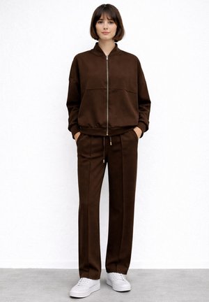 Kobieta stojąca przy białej ścianie, ubrana w brązową kurtkę zapinaną na zamek, dopasowane brązowe spodnie oraz białe trampki, z rękami w kieszeniach.