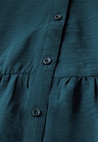 Chemise bleu sarcelle à boutons avec un tissu texturé, cinq boutons sur le devant, et un design froncé à la taille.