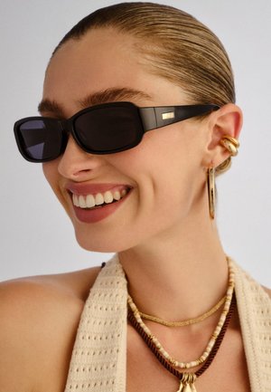 Žena sa zalizanim kosom koja nosi crne pravokutne sunčane naočale, zlatne obruče naušnice, slojevite ogrlice od perlica i bež pleteni top, smiješi se.