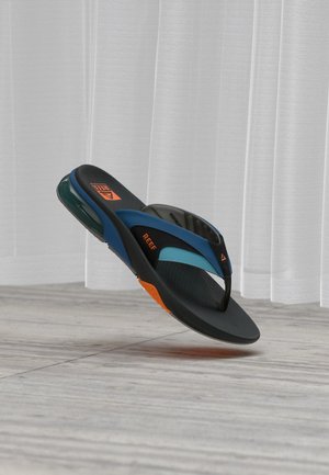 Μαύρο και μπλε σανδάλι τύπου flip-flop Reef με πορτοκαλί λεπτομέρειες, που αιωρείται πάνω από ξύλινο πάτωμα με λευκές κουρτίνες στο φόντο.