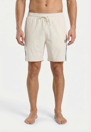 Uomo che indossa pantaloncini beige con coulisse e logo sulla coscia sinistra, in piedi a piedi nudi contro uno sfondo bianco semplice.