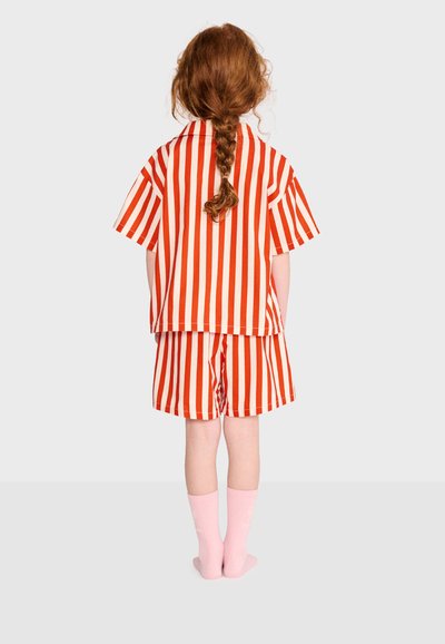 Enfant aux cheveux roux tressés portant une chemise à rayures verticales rouges et blanches ainsi qu'un short, debout pieds nus sur un fond uni.