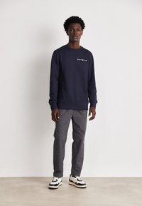 Námornícky crewneck sveter s bielym logom "GANT 1949", kombinovaný so sivými strihovými nohavicami a čierno-bielymi teniskami. Hladká textúra látky.