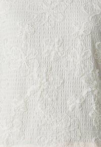 Top de tela blanca texturizada con aplicaciones florales en relieve. El diseño incluye una superficie arrugada con intrincados patrones de flores.