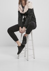 Giacca a vento nera e rosa con cappuccio, colletto alto e polsini elastici, abbinata a pantaloni tapered neri e sneakers nere su uno sgabello bianco.