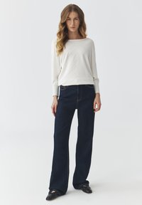 Hvid langærmet sweater med zigzag mønster, parret med mørkeblå høj talje flared jeans og sorte flade sko. Neutral baggrund.