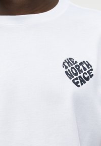 Κοντινή λήψη λευκού T-shirt με μαύρο λογότυπο σε σχήμα καρδιάς που γράφει "THE NORTH FACE" στο αριστερό στήθος.