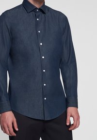 Mann in einem dunkelblauen Langarmhemd mit Knöpfen und schwarzen Hosen, der vor einem schlichten grauen Hintergrund steht.