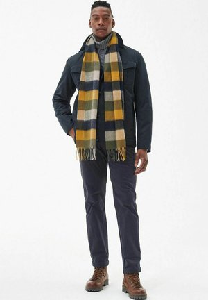 Herenjas in marineblauw over een grijze coltrui, gecombineerd met een wollen sjaal met een multicolor geruit patroon, donkere broek en bruine veterlaarzen.