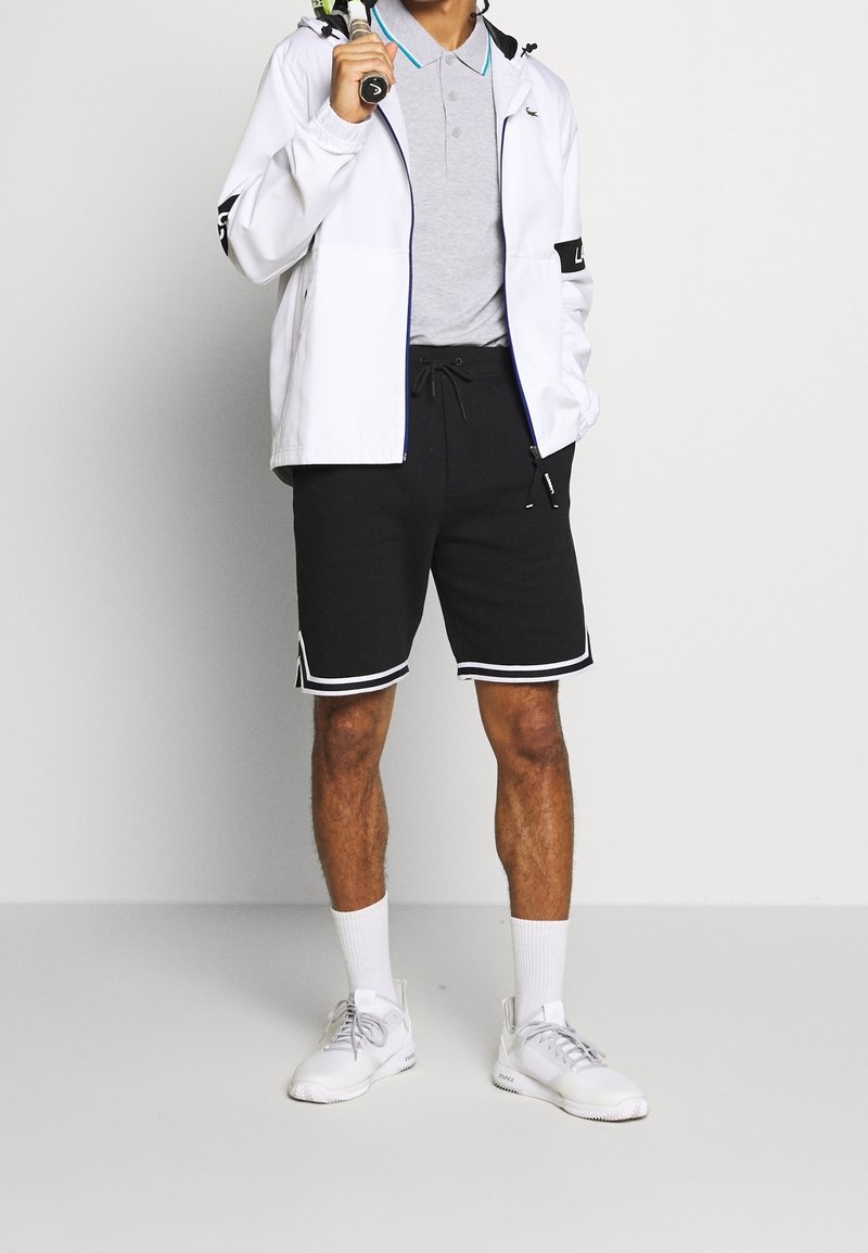 Vit zip-jacka med svarta detaljer, grå polo, svarta shorts med vita kanter och vita sportskor med grå detaljer.