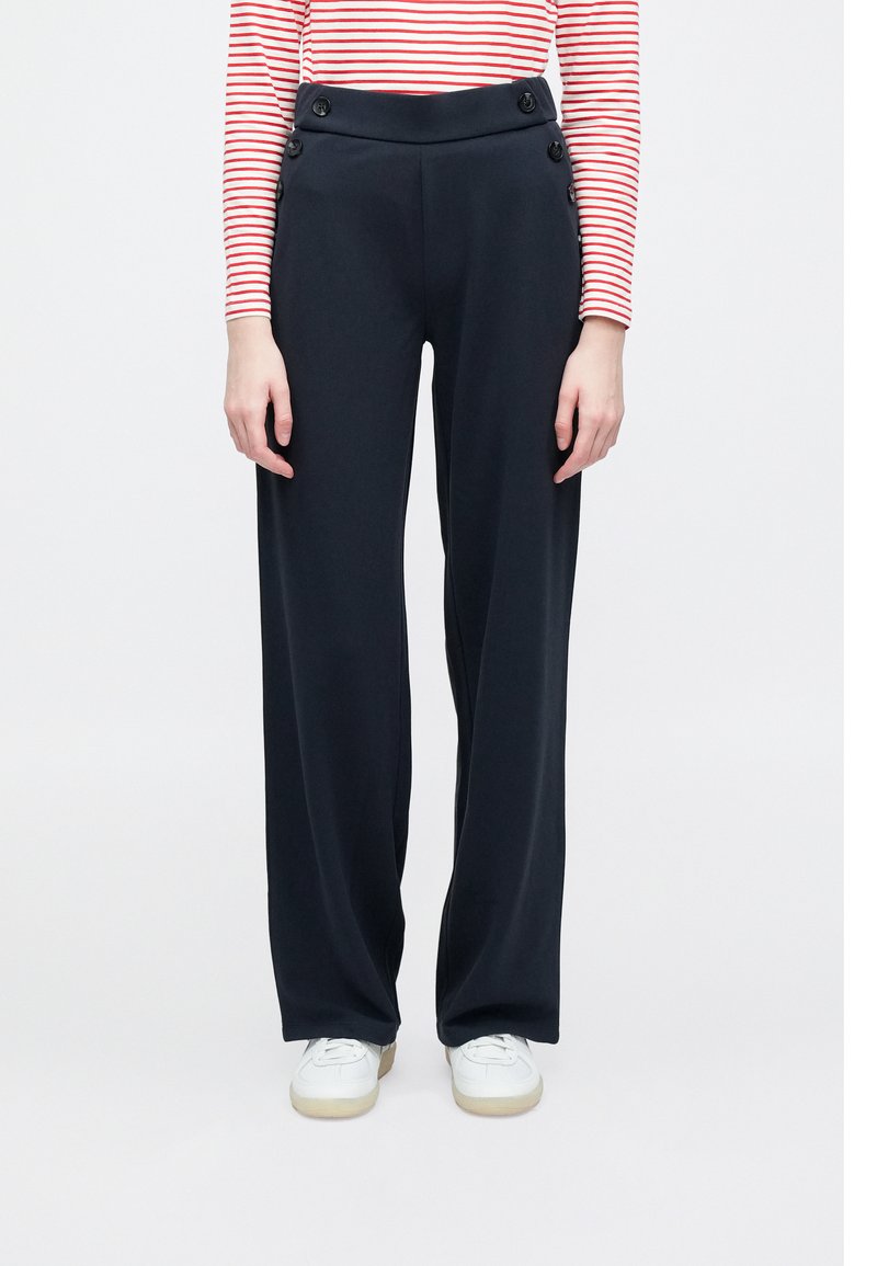 Granatowe spodnie z wysokim stanem o luźnym kroju, z bocznymi guzikami i gładką fakturą, zestawione z białymi sneakersami.