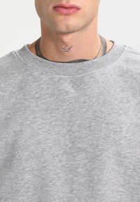 Sudadera gris de cuello redondo hecha de una mezcla de algodón texturizado, con un cuello acanalado y un tatuaje visible en la zona del cuello.