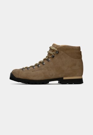 Brune suède tursko med metalløyer, beige snører, polstret ankelkrage og svart gummisåle med knopper vist i profil.