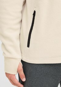 Lyst beige fleecejakke med lynlås-lomme i siden og ribbede manchetter, der har en glat tekstur og tætsiddende design.