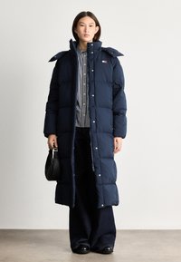 Dlhá námornícka pufrová bunda s veľkým golierom a cvokmi, prešívaná textura, skombinovaná so pruhovanou košeľou a tmavými širokými džínami.