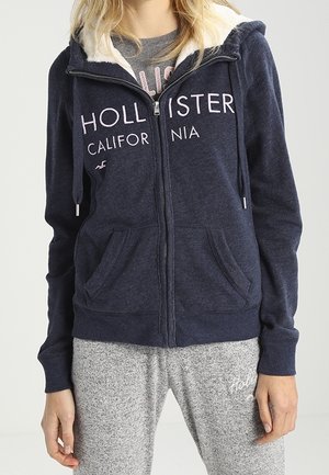 Vrouw draagt een marineblauwe Hollister vest met rits, voorzien van een witte fleece voering, en grijze joggingbroek met Hollister-logo op de linker dij.