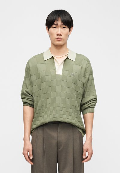 Νεαρός άνδρας φορώντας πράσινο καρό πουλόβερ με γυρισμένα μανίκια, κρεμ γιακά πόλο, μαργαριταρένιο κολιέ και καφέ πλισέ παντελόνι σε απλό φόντο.