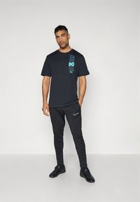 Black t-shirt with turquoise "Just Do It" text, paired with black tapered pants featuring a small Nike logo. Model wears black athletic shoes.
