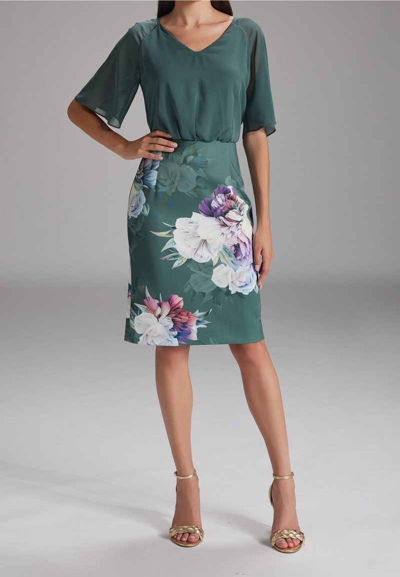 Femme portant une robe verte à manches flottantes avec un grand imprimé floral, associée à des sandales à talons hauts ouvertes dorées, debout devant un fond uni.