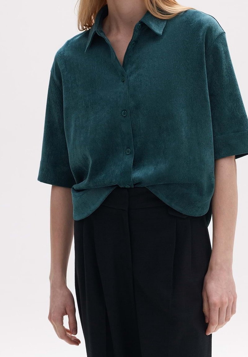 Femme portant une chemise boutonnée à manches courtes vert foncé rentrée dans un pantalon noir taille haute, montrée du cou jusqu'aux hanches.