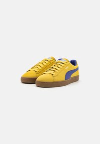 Yellow suede sneakers with a purple accent stripe, flat laces, and a brown rubber sole. Features a padded collar and logo on the tongue.