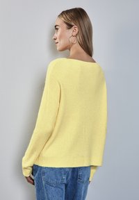 Gul ribbad tröja med avslappnad passform och bred halsringning, kombinerad med ljusa denimjeans. Har långa ärmar och textilstruktur.