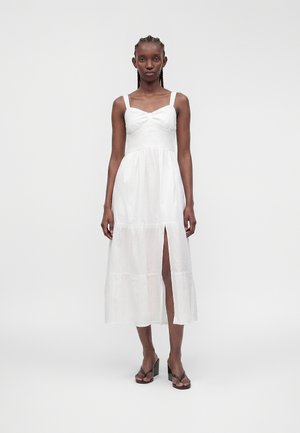 Femme aux cheveux tressés portant une robe midi blanche sans manches avec une fente à l'avant et des sandales à talons noires, debout sur un fond uni.