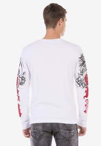 Weißes Langarmshirt mit detaillierter schwarzer Kunst auf den Ärmeln und roten Schriftzügen. Weiche Baumwolle mit normalem Schnitt und Rundhalsausschnitt.