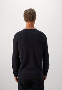 Bărbat cu păr scurt îmbrăcat într-o cămașă lungă neagră și pantaloni negri, surprins din spate pe un fond simplu deschis.
