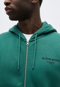 Férfi zöld cipzáras kapucnis felsőben, húzózsinórokkal, a mellkasán "BJÖRN BORG STOCKHOLM 1956" felirattal, fehér ing alatt.