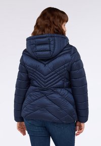 Donna con capelli castani che indossa una giacca a vento blu navy trapuntata con cappuccio e jeans blu, di spalle su uno sfondo bianco.