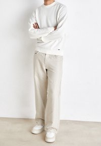 Felpa bianca a maglia testurizzata con polsini a costine, abbinata a pantaloni palazzo beige chiaro e sneakers bianche, in piedi davanti a uno sfondo neutro.