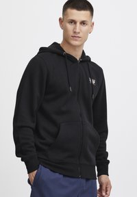 Zwarte zip-up hoodie gemaakt van een zachte katoenmix. Bevat twee voorzakken, verstelbare trekkoorden en een klein logo op de borst.