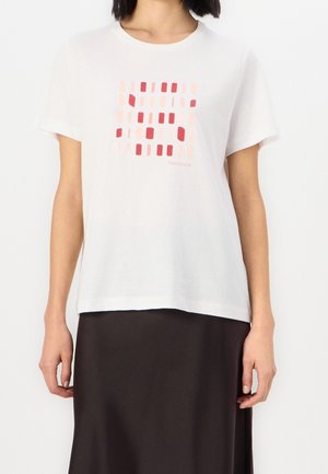 Kobieta ubrana w białą koszulkę z krótkim rękawem z geometrycznym wzorem kwadratów w kolorze różowym i czerwonym oraz czarną spódnicę.