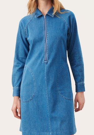 Frau trägt ein langärmeliges blaues Jeanskleid mit Reißverschluss vorne und großen runden Taschen vor hellem Hintergrund.