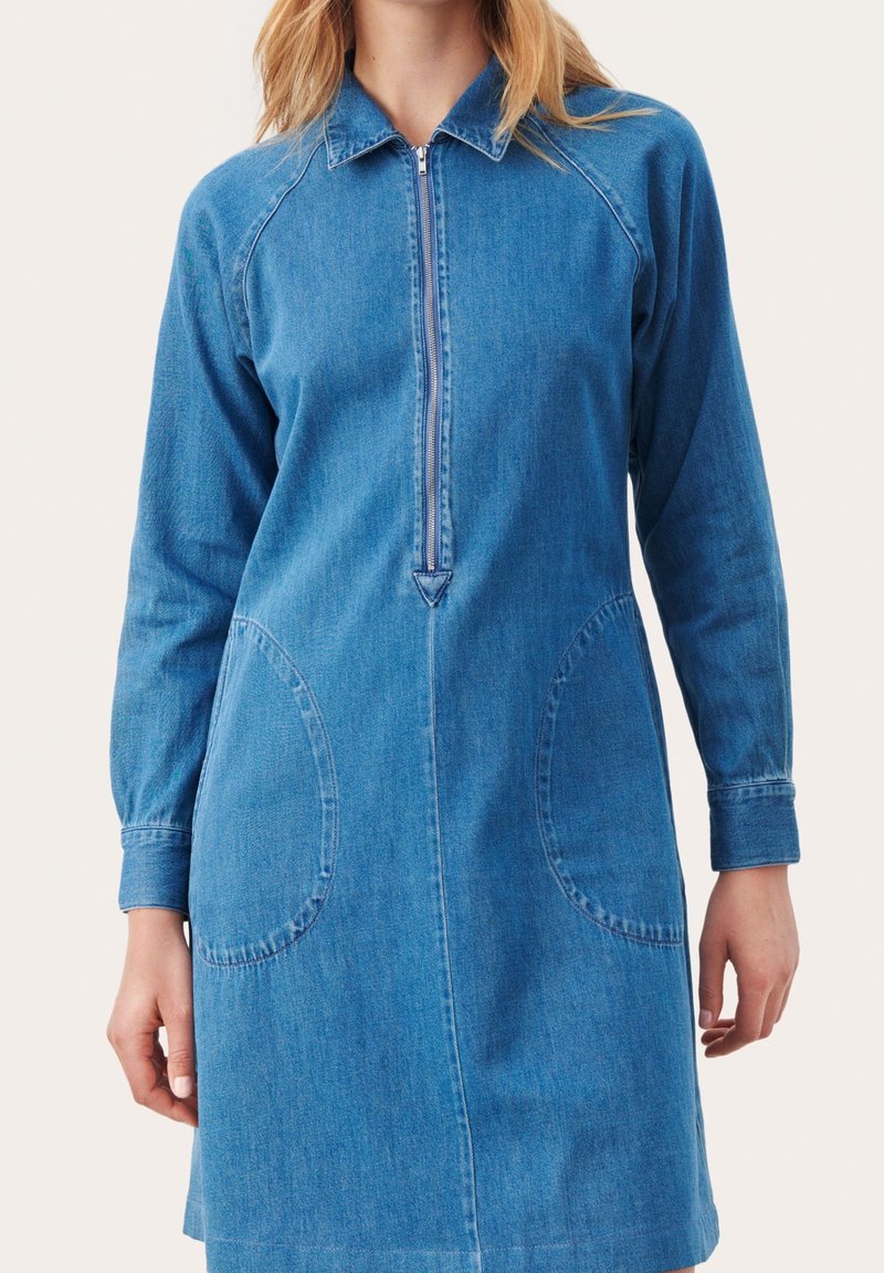 Femme portant une robe en denim bleu à manches longues avec une fermeture éclair devant et de grandes poches rondes sur un fond clair.