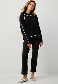 Maglione nero a maglia con dettagli bianchi, vestibilità comoda, polsini a costine, abbinato a pantaloni neri attillati e ballerine grigio chiaro. Sfondo neutro.