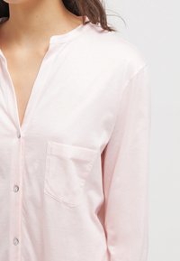 Camisa de manga larga de color rosa claro con botones en la parte delantera, diseño sin cuello y un único bolsillo en el pecho. Tejido de algodón suave con textura lisa.