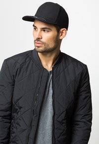 Schwarze gesteppte Bomberjacke mit einem Reißverschluss, über einem grauen T-Shirt getragen, kombiniert mit einer schwarzen Flachschirmmütze. Saubere Linien und minimale Akzente.