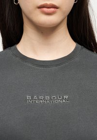 Κοντινό πλάνο ενός ατόμου που φοράει σκοτεινό γκρι φούτερ Barbour International με κεντημένο κείμενο στο στήθος.