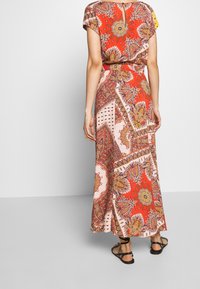 Robe maxi en tissu léger, présentant un motif paisley rouge et rose, des manches courtes, une ceinture à la taille et des fentes sur les côtés. Sandales noires visibles.