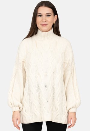 Junge Frau mit langen dunklen Haaren, die goldene Creolen und einen weißen Strickpullover mit Rollkragen und ballonförmigen Ärmeln trägt.