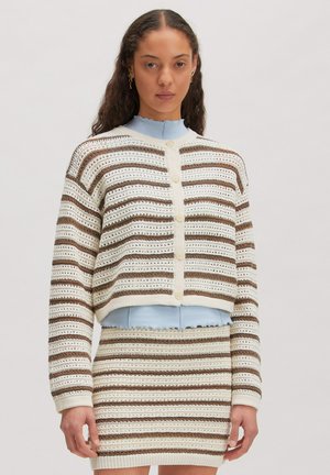 Vrouw draagt een crème, bruin en wit gestreepte gebreide cardigan en bijpassende rok over een lichtblauw topje, staand tegen een effen achtergrond.