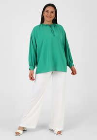 Grüne Bluse mit rundem Halsausschnitt und Bindedetail, langen Ballonärmeln, getragen mit weißen weitgeschnittenen Hosen und Absatzsandalen.